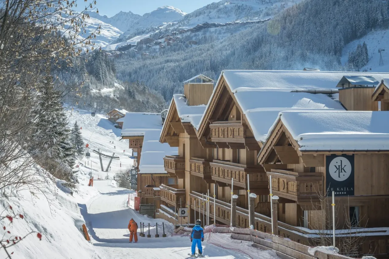 Vue sur des chalets enneigés au bord d'une piste de ski avec des skieurs en arrière-plan - Séjour au Ski en Savoie - Le Kaïla
